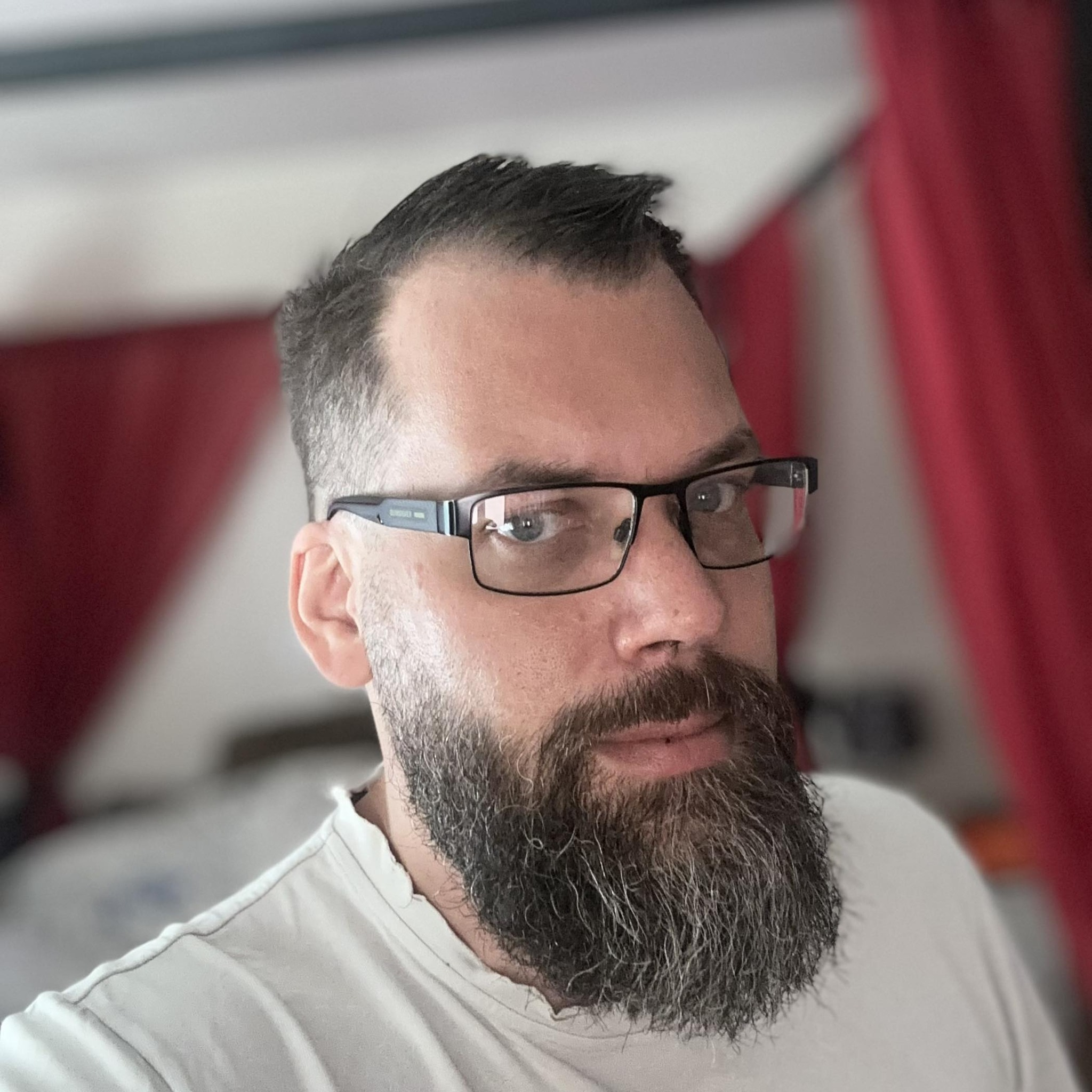 A close-up image of a man with a short haircut, black-framed glasses, and a white t-shirt. He has a thick, dark beard and ...